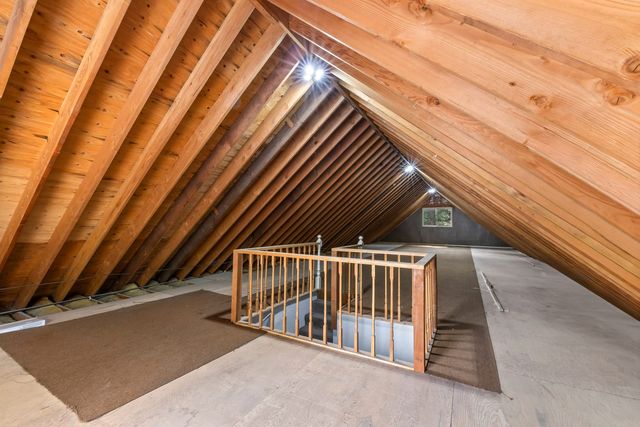 Unfinished attic in a Wrightwood home with exposed wood rafters, a railed stairwell opening, carpet runners over plywood flooring and a small gable window