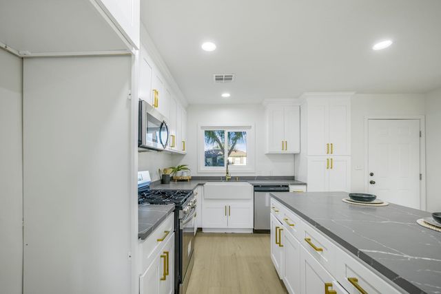 Empty renovated kitchen in a Pomona house with white cabinets, brass handles, gray stone counters, a farmhouse sink and a gas range
