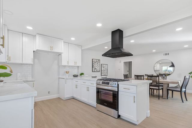 Renovated kitchen in a Pomona house with white shaker cabinets, brass pulls, a black hood over a gas range and a dining table beside the peninsula