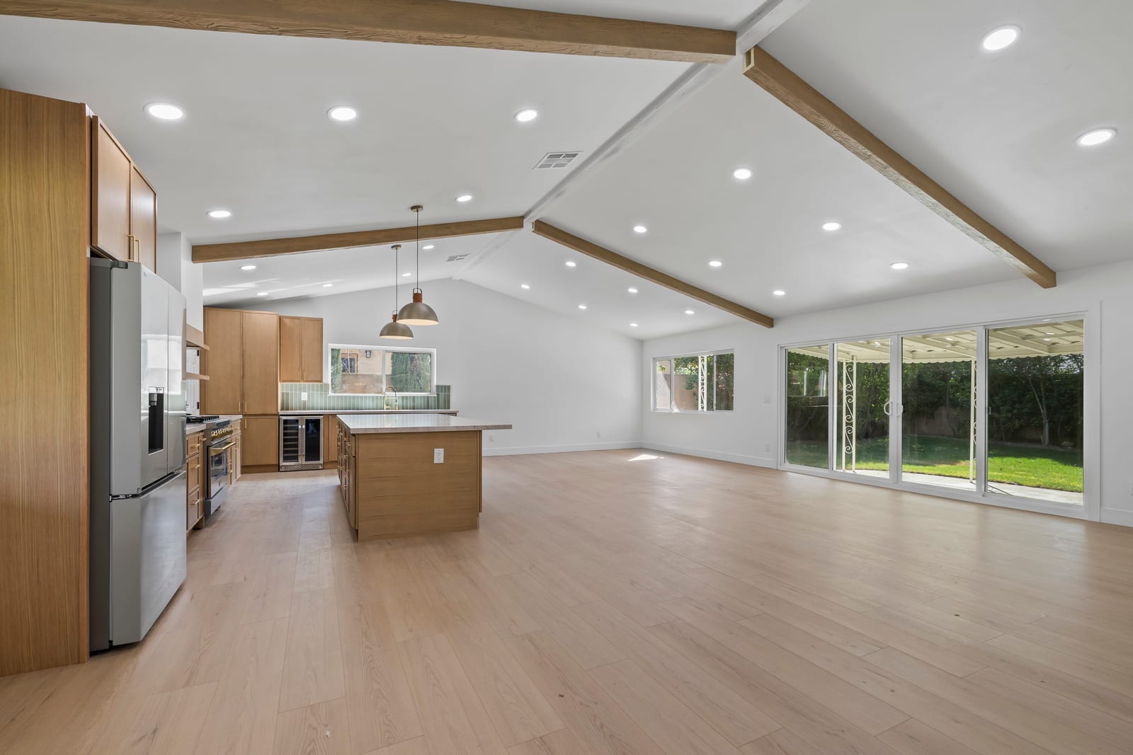 Empty open-plan kitchen and family room with wood ceiling beams, an island under two pendant lights, light plank flooring and sliding glass doors to a covered patio and lawn